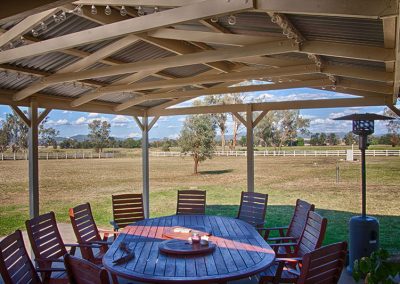 Covered Outdoor Dining area - natural wood setting - with outdoor gas heater to the right. The view from this lovely outdoor setting is a large open paddock area with a white post and rail fencing and trees in the background.