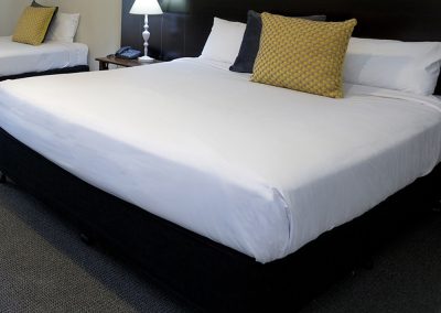 Queen size bed covered with white linen and beautiful textured gold and black cushions. On the side table is a room telephone and a white lampstand