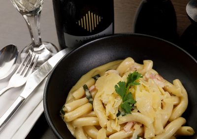 A pasta dish with cheese dished up on a black plate and a bottle of wine in the background