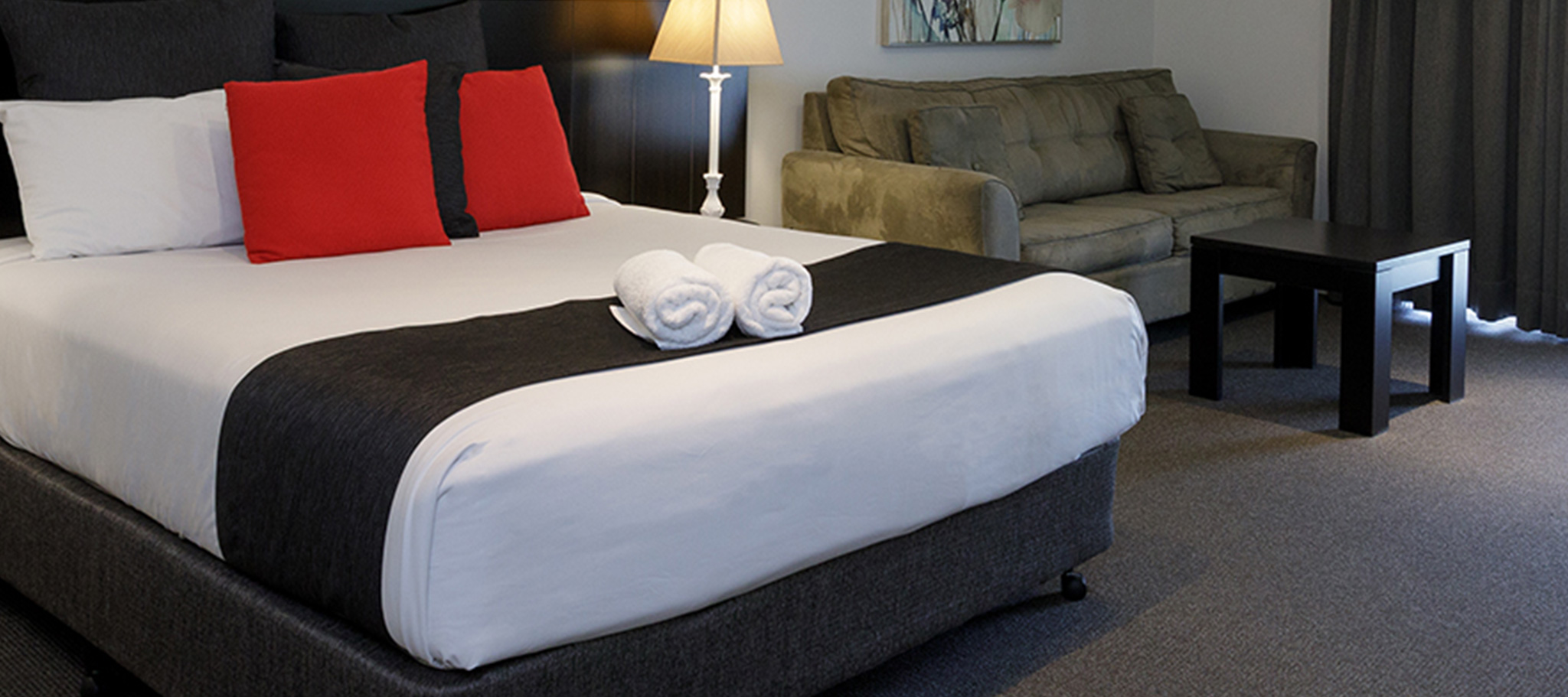 A queen size bed with beautiful white linen covers and red scatter cushions. Two rolled up white towel are displayed on the edge of the bed. There is a couch and table in the room and the night stand is on, making it very homely and inviting.