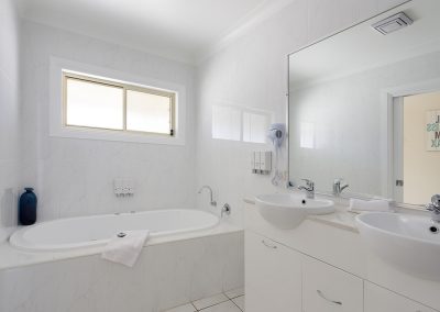 Stockmans Motel Spa bathroom. all white with two basins. A blue bottle is in the corner on the spa rim for decoration and a pure white towel is folded and draped over the edge of the bath.